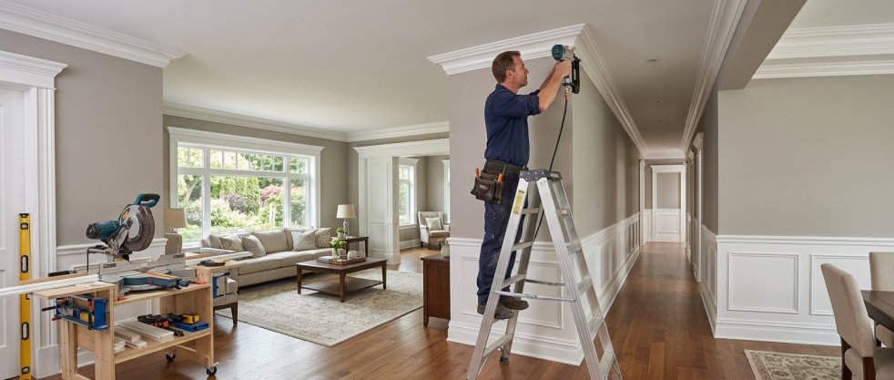 Contractor on ladder installing crown molding and wall trim in a bright finished home interior